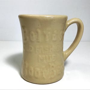 May include: A beige ceramic mug with a curved handle. The mug has embossed text that reads "Old Fashioned Mug". The mug is a light tan color and has a smooth finish. The mug is suitable for hot or cold beverages.