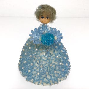 May include: A vintage doll with a light blue beaded dress and a matching floral bodice. The doll has a small face with blue eyes and gray hair. The dress is adorned with small white pearls.