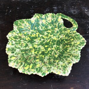 May include: A green and yellow ceramic leaf-shaped serving dish with a handle. The dish has a speckled glaze with green, yellow, and brown spots.