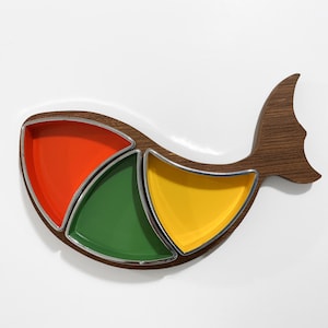 May include: A fish-shaped serving tray crafted from dark brown wood with a prominent tail fin. The tray features three separate, removable, triangular-shaped bowls with smooth, glossy interiors in vibrant orange, forest green, and bright yellow.