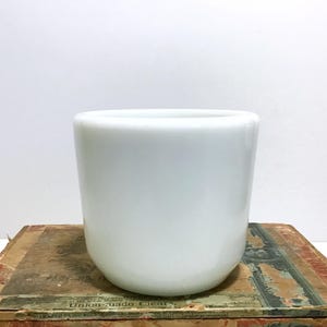 May include: A white, cylindrical ceramic planter with a smooth, glossy finish. The planter has a rounded base and a wide opening. It is sitting on a vintage cigar box.