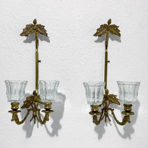 May include: A pair of wall-mounted candle sconces made of brass with a leaf-shaped top ornament. Each sconce features two arms holding clear, fluted glass candle cups. The brass metalwork includes detailed leaf motifs at the base of the cups.