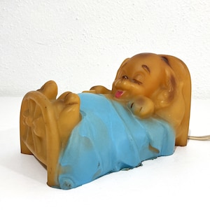 May include: A molded plastic lamp shaped like a cartoon dog sleeping in a small bed. The dog is tan with closed eyes and a small pink tongue visible. It is covered by a bright blue blanket that drapes over the bed frame, which has a wheel design.