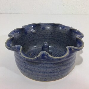 May include: A handmade, blue ceramic ashtray with a ruffled edge and a central cone-shaped rest. The ashtray has a speckled texture and a rustic, artisanal appearance. The rim is a lighter shade of blue.