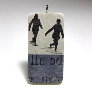 May include: A rectangular pendant with a silver-colored loop at the top. The top portion features a black and white image of two people walking in the snow. The bottom portion has a dark blue background with white text. The pendant is rectangular.