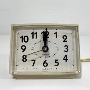 May include: A rectangular Westclox electric alarm clock with a cream-colored plastic casing and a clear front cover. The white clock face features bold black numerals from 1 to 12, black hour and minute hands, a thin gold second hand, and text.