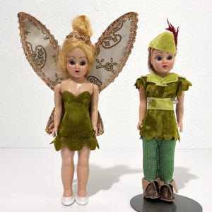May include: Two vintage dolls, one dressed as a fairy with gold-trimmed wings and a green dress, the other as Peter Pan in a green outfit and hat. The fairy doll has blonde hair and white shoes. The Peter Pan doll has brown shoes.