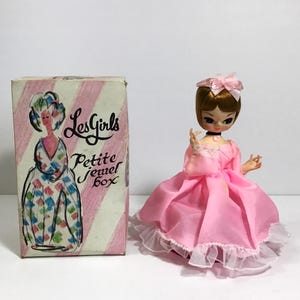 May include: A pink and white doll with a pink dress and a white lace trim. The doll has brown hair and blue eyes. The doll is sitting on a white base. The doll is in a box with the words "Les Girls Petite Jewel Box" on it.