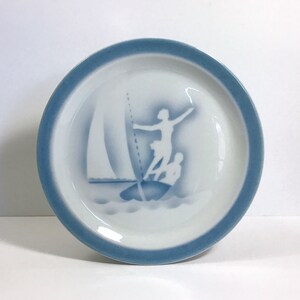 May include: A white ceramic plate with a blue rim and a blue and white silhouette design of a person sailing a sailboat.