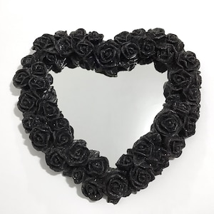 May include: A heart-shaped mirror framed with black roses. The roses have a glittery finish, and the mirror reflects the surrounding environment. The design is set against a white background, highlighting the contrast between the dark roses and the reflective surface.