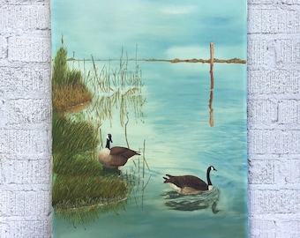 May include: An oil painting of a serene lake scene with two Canada geese. One goose stands on the grassy bank, while the other swims in the water. The painting features a muted color palette of blues, greens, and browns, with a wooden post in the distance.