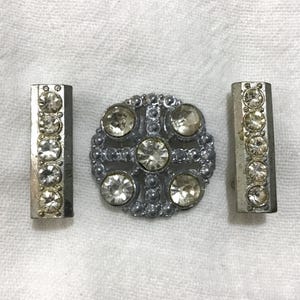 May include: A set of three silver-toned metal embellishments. The set includes two rectangular bars and a circular, cross-shaped piece, all adorned with clear, sparkling rhinestones. The pieces are likely for clothing or craft projects.