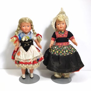May include: Two vintage dolls wearing traditional Dutch clothing. The doll on the left has blonde hair and is wearing a red, white, and blue dress with a black bodice. The doll on the right has blonde hair and is wearing a black dress with a red, white, and blue bodice and a white lace cap.