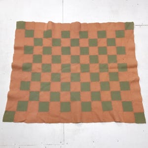May include: A rectangular blanket with a checkerboard pattern. The blanket features alternating squares of orange and olive green. The blanket is made of a soft material and has a stitched border.