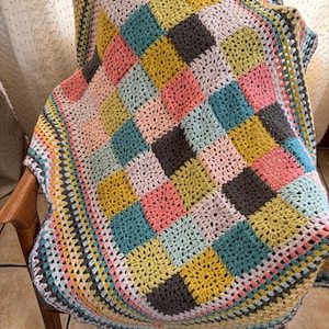 Può includere: Una coperta all'uncinetto colorata con un design a patchwork in tonalità di rosa, giallo, verde, blu e grigio. La coperta ha un bordo all'uncinetto in una varietà di colori.