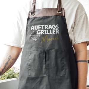 May include: A dark gray apron with brown leather straps and a pocket. The apron has the text "AUFTRAGS GRILLER Marcel" in white and gold. The apron is designed for grilling.