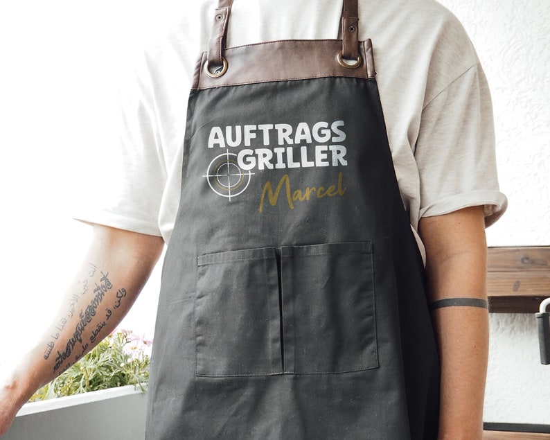 May include: A dark gray apron with brown leather straps and a pocket. The apron has the text "AUFTRAGS GRILLER Marcel" in white and gold. The apron is designed for grilling.