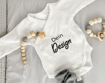 Könnte beinhalten: Ein weißer Langarm-Babybody mit dem schwarzen Schriftzug "Dein Design" auf der Vorderseite. Das Set enthält eine Schnullerkette mit Holz- und Häkelperlen sowie ein Paar graue und weiße Socken mit strukturiertem Strickmuster.
