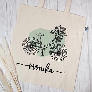 Könnte beinhalten: Eine weiße Stofftasche mit einer schwarz-weißen Illustration eines Fahrrads mit einem Blumenkorb. Der Text "monika" ist in Kursivschrift unter dem Fahrrad geschrieben.
