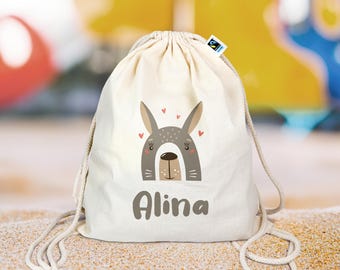 Op de afbeelding: Een witte trekkoordtas met een schattig cartoon konijnengezicht en de naam "Alina" erop gedrukt. De tas heeft een bruin en grijs konijnengezicht met roze hartjes eromheen.