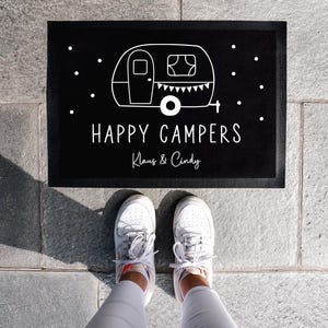 Könnte beinhalten: Schwarze Willkommensmatte mit weißem Text, der "Happy Campers Klaus & Cindy" lautet. Ein weißer Cartoon-Wohnwagen befindet sich über dem Text.