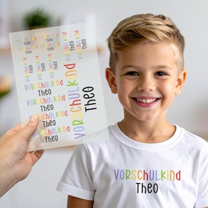 Peut inclure: Un t-shirt blanc avec le mot "VORSCHULKIND" en couleurs arc-en-ciel et le nom "Theo" en noir. Une feuille d'autocollants avec le même texte est tenue. Un jeune garçon sourit à la caméra.