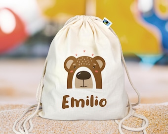 Op de afbeelding: Een witte trekkoordtas met een bruin beren gezicht en de naam "Emilio" erop gedrukt. De tas heeft een wit trekkoord en is gemaakt van natuurlijk katoen.