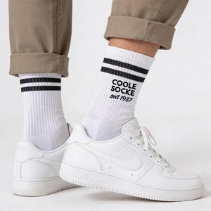 Könnte beinhalten: Weiße Socken mit zwei schwarzen Streifen und dem Text "COOLE SOCKE seit 1987". Die Socken werden mit weißen Turnschuhen und einer khakifarbenen Hose getragen. Die Turnschuhe haben einen weißen Nike-Swoosh.