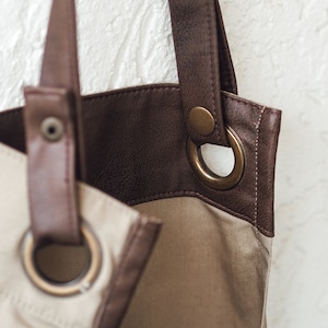 May include: Close-up of a beige and brown tote bag. The bag features a light beige canvas body with brown leather accents and straps. Large metal eyelets and a snap closure are visible. The bag has a simple, classic design.