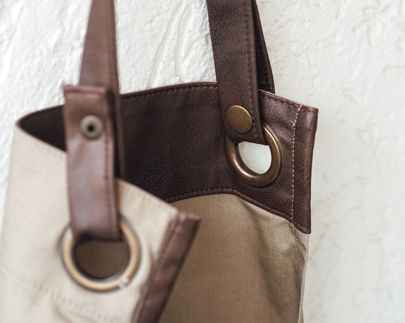 May include: Close-up of a beige and brown tote bag. The bag features a light beige canvas body with brown leather accents and straps. Large metal eyelets and a snap closure are visible. The bag has a simple, classic design.