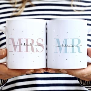 Könnte beinhalten: Zwei weiße Keramikbecher mit schwarzen Henkeln. Der Becher links sagt "MRS Schmitz" in rosa Schrift mit kleinen weißen Punkten. Der Becher rechts sagt "MR Schmitz" in blauer Schrift mit kleinen weißen Punkten.