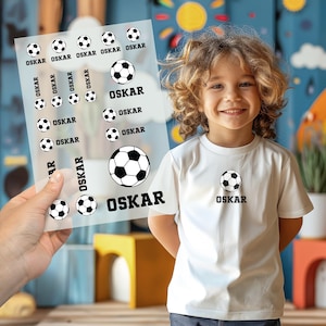 Peut inclure: Un enfant porte un t-shirt blanc avec un ballon de football et le nom "OSKAR". Une main tient une feuille d'autocollants avec des motifs de ballons de football et le nom "OSKAR". Les autocollants sont en noir et blanc.