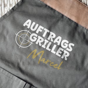 May include: Gray apron with brown leather accents and the text "AUFTRAGS GRILLER Marcel" in white and gold. Features a target graphic. Includes a pocket and metal grommets.