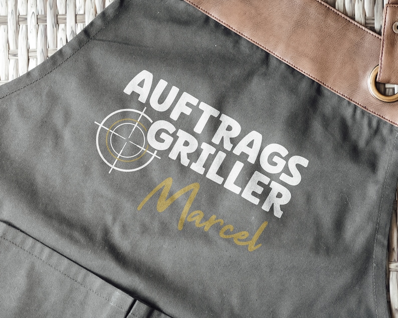 May include: Gray apron with brown leather accents and the text "AUFTRAGS GRILLER Marcel" in white and gold. Features a target graphic. Includes a pocket and metal grommets.