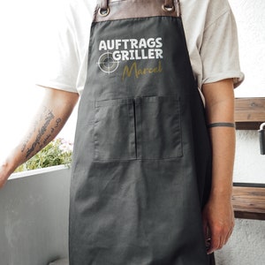 May include: A black apron with brown leather straps and a brown leather neck piece. The apron has two front pockets and the text "AUFTRAGS GRILLER Marcel" printed in white and gold.