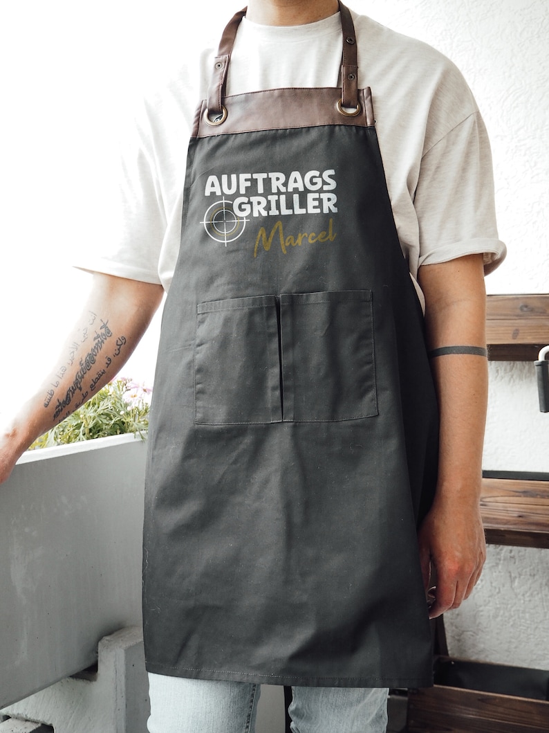May include: A black apron with brown leather straps and a brown leather neck piece. The apron has two front pockets and the text "AUFTRAGS GRILLER Marcel" printed in white and gold.