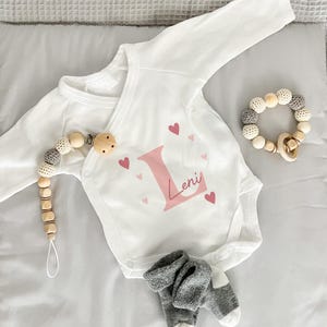 Könnte beinhalten: Ein weißer Langarm-Babybody mit einem großen rosa Buchstaben L, dem Namen Leni in Schreibschrift und kleinen rosa Herzen. Dazu gehören ein Schnullerhalter aus Holzperlen, eine Holzperlen-Rassel und ein Paar graue Babysocken.