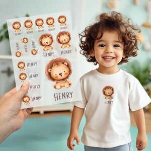 Può includere: Una maglietta bianca con una grafica di leone e il nome "HENRY" stampato sotto. Un foglio di adesivi con lo stesso disegno di leone e il nome "HENRY" è tenuto in mano. Gli adesivi e la maglietta presentano un simpatico leone dei cartoni animati.