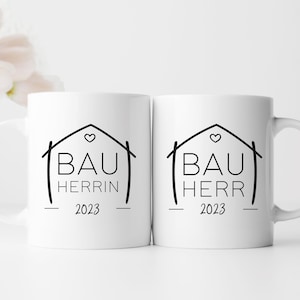 Könnte beinhalten: Zwei weiße Keramikbecher mit schwarzem Text und einer Hausumrandung. Der Text auf den Bechern lautet "Bauherrin 2023" und "Bauherr 2023".