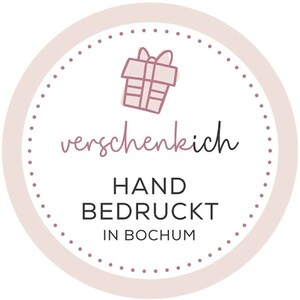 May include: A round, light pink and white sticker with a dotted border. The sticker features a line drawing of a gift box and the text "verschenkich", "HAND BEDRUCKT IN BOCHUM" in a mix of script and block letters.