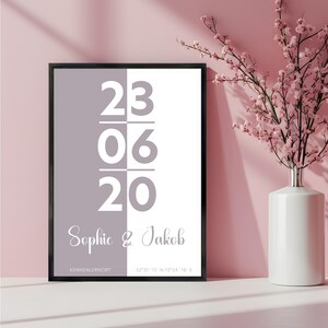 Peut inclure: Un cadre avec un fond divisé en gris et blanc. L'impression affiche la date 23-06-20, les noms "Sophie & Jakob", ainsi que le lieu et les coordonnées. Un vase blanc avec des fleurs roses est à droite.