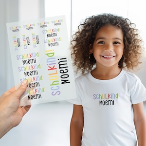 Può includere: Una bambina sorride, indossa una maglietta bianca con la scritta "SCHULKIND! Noemi" in lettere colorate. Un foglio di adesivi con lo stesso testo è tenuto vicino. Gli adesivi e la maglietta sono probabilmente per la scuola.