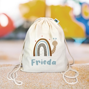 Könnte beinhalten: Eine weiße Kordeltasche mit einem Regenbogen- und Schneckenmotiv. Die Tasche ist mit dem Namen "Frieda" bedruckt.