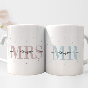 Könnte beinhalten: Zwei weiße Keramikbecher mit herzförmigen Griffen. Der Becher links sagt "MRS Kruger" in rosa Schrift mit grauen Punkten. Der Becher rechts sagt "MR Kruger" in blauer Schrift mit grauen Punkten.
