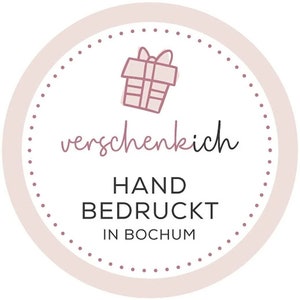 May include: A circular sticker with a light pink border and a dotted inner circle. The sticker features a hand-drawn gift box illustration and the text "verschenkich", "HAND BEDRUCKT IN BOCHUM" in a mix of script and block letters.
