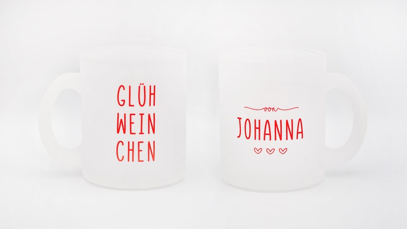 May include: Two frosted glass mugs with handles. One mug has the red text "GL&Uuml;H WEIN CHEN", and the other has "von JOHANNA" with three hearts. The mugs are white and sit on a white background.