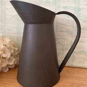 May include: A dark grey metal jug with a curved handle. The jug has a wide spout and a tapered body. The handle is arched and attached to the side of the jug. The jug is sitting on a wooden surface.