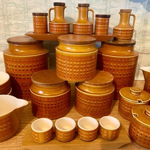May include: A collection of vintage kitchen canisters and serving pieces in shades of brown and orange. The set includes jars, cups, and jugs with wooden lids and decorative dot patterns. The word "TEA" and "COFFEE" are visible on some of the jars.