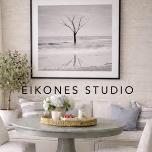 Peut inclure: Une photographie encadrée en noir et blanc d'un arbre dans l'océan est suspendue au-dessus d'une table ronde grise. La table est ornée d'un plateau tressé, de fleurs et de petits plats. Le texte "EIKONES STUDIO" est en dessous.