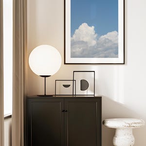 Könnte beinhalten: Moderne Inneneinrichtung mit einem schwarzen Schrank, einer kugelförmigen weißen Lampe und gerahmter Kunst. Das Kunstwerk umfasst einen gerahmten Druck eines blauen Himmels mit flauschigen weißen Wolken. Ein kleiner, strukturierter Beistelltisch vervollständigt die Szene.